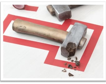 May include: Two hammers with silver heads and bronze handles are on a white surface with a red and white geometric design. One hammer has a broken head with chocolate pieces.