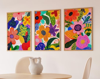 May include: Three framed abstract floral prints with a colourful, whimsical design. The prints feature a variety of flowers, leaves, and geometric shapes in bright colours like pink, yellow, orange, green, and blue. The prints are hung on a white wall above a wooden table with two white chairs.