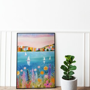 May include: A framed painting of a colorful cityscape with sailboats on a blue body of water. The water is sparkling and there are flowers in the foreground. The sky is a soft pink and purple.