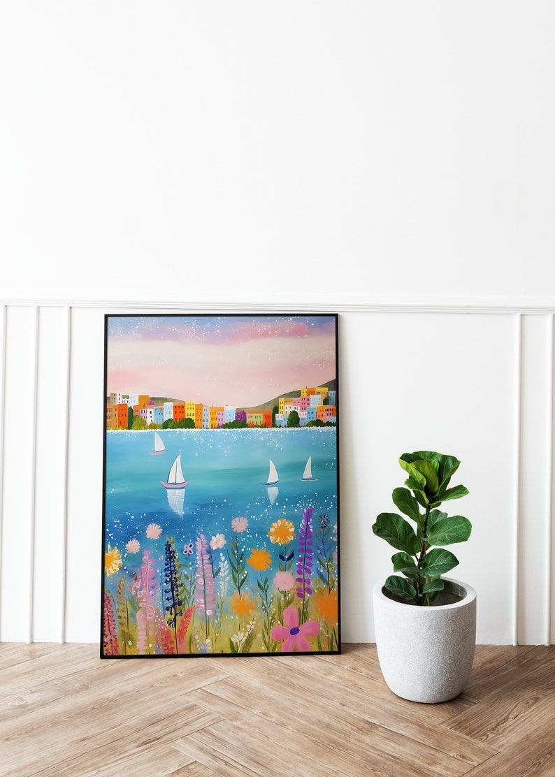 May include: A framed painting of a colorful cityscape with sailboats on a blue body of water. The water is sparkling and there are flowers in the foreground. The sky is a soft pink and purple.