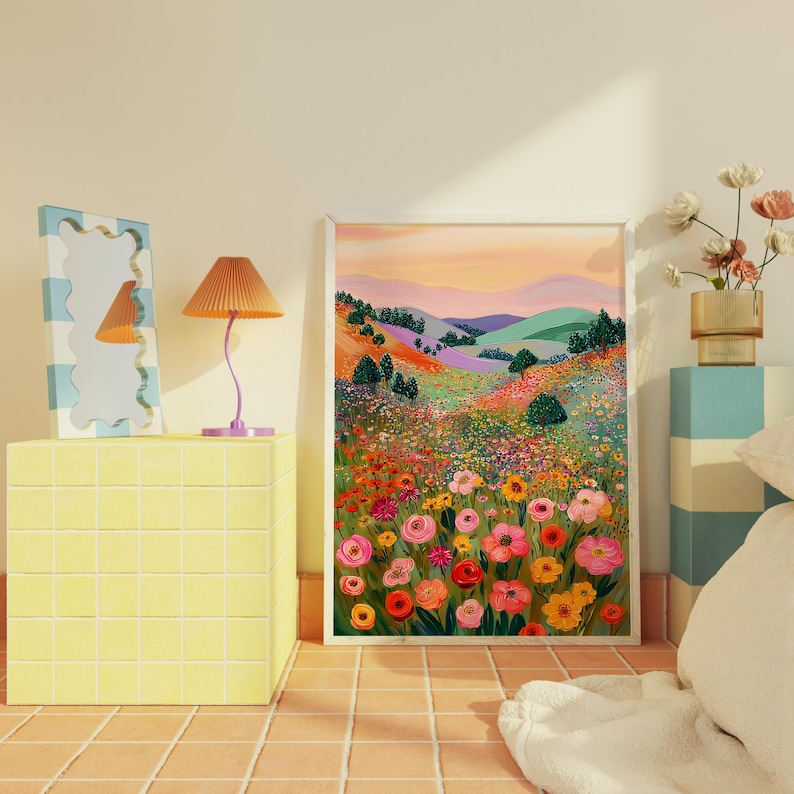 May include: A framed art print depicts a vibrant, colorful landscape with rolling hills and a field of flowers. The artwork features a palette of pinks, oranges, greens, and purples. A yellow side table, a wavy mirror, and a lamp are also in the image.