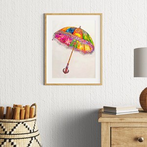 Op de afbeelding: Een ingelijst aquarel schilderij van een kleurrijke, sierlijke paraplu met een houten handvat. De paraplu is geschilderd in tinten roze, geel, groen, blauw en paars. De achtergrond is wit.