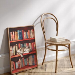 May include: A framed print of a cat sleeping on a bookshelf filled with colorful books. The bookshelf is red and the cat is white and gray. The print is hanging on a white wall next to a wooden chair.