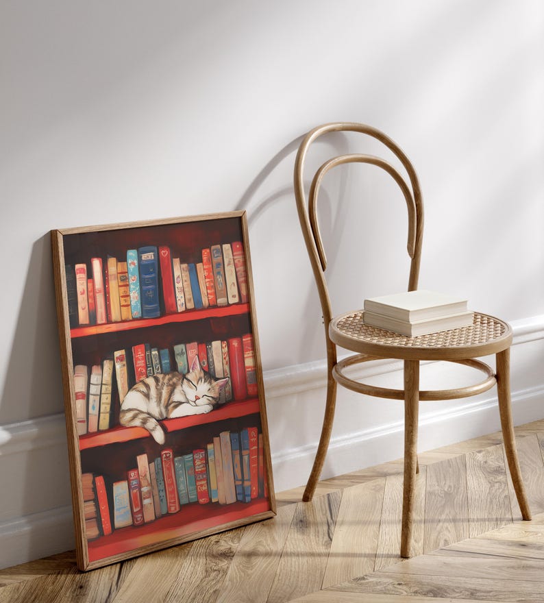 May include: A framed print of a cat sleeping on a bookshelf filled with colorful books. The bookshelf is red and the cat is white and gray. The print is hanging on a white wall next to a wooden chair.