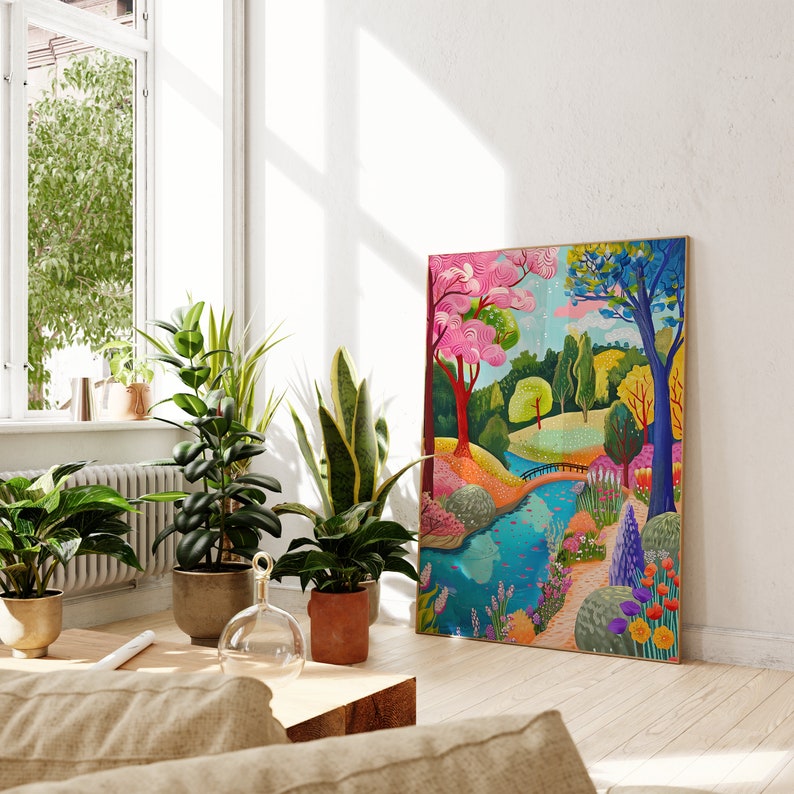 May include: A colorful framed art print depicting a vibrant landscape scene with a river, bridge, and trees. The artwork features a pink tree, blue tree, and various other trees and flowers. The print is leaning against a white wall, with potted plants in the foreground.