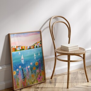 May include: A framed painting of a colorful cityscape with sailboats on a blue body of water. The painting is surrounded by a wooden frame and is leaning against a white wall. The painting is in front of a wooden chair with a woven seat.