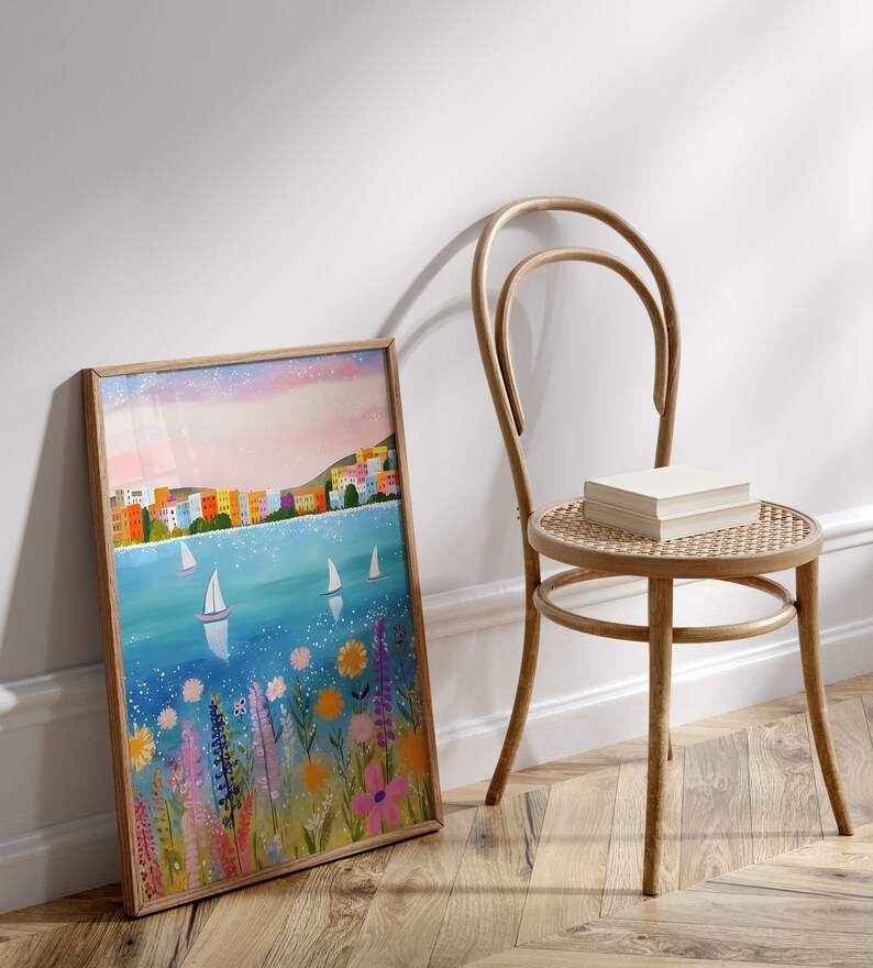 May include: A framed painting of a colorful cityscape with sailboats on a blue body of water. The painting is surrounded by a wooden frame and is leaning against a white wall. The painting is in front of a wooden chair with a woven seat.