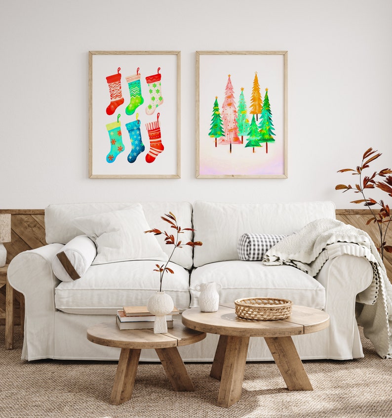 Puede incluir: Dos pinturas de acuarela enmarcadas en una pared blanca sobre un sof&aacute; blanco con dos mesas de caf&eacute; de madera. La pintura de la izquierda presenta seis calcetines de Navidad coloridos. La pintura de la derecha presenta un grupo de cinco &aacute;rboles de Navidad coloridos.