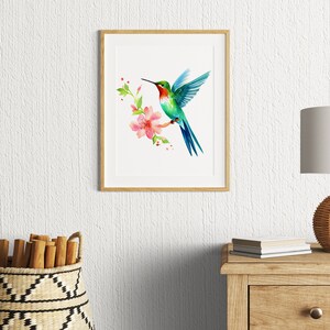Puede incluir: Una pintura de acuarela enmarcada de un colibr&iacute; en vuelo, con una flor rosa y hojas verdes. El colibr&iacute; es azul, verde y rojo.