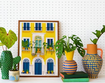 May include: A framed illustration of a yellow building with blue windows and a balcony. The building is surrounded by potted plants. The illustration is on a white background with black polka dots.