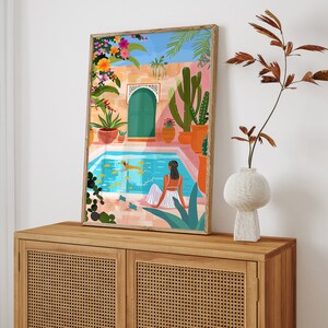 May include: A colorful illustration of a woman relaxing by a pool in a Moroccan-style courtyard. The pool is surrounded by lush greenery, including cacti and palm trees. The woman is wearing a white dress and has her hair pulled back. The illustration is framed in a wooden frame.