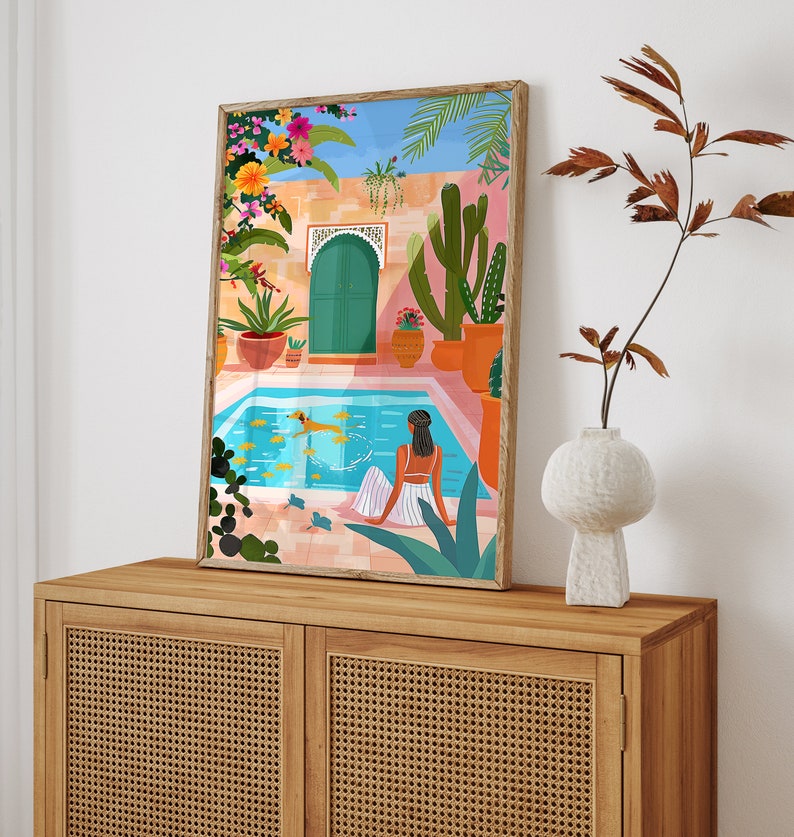 May include: A colorful illustration of a woman relaxing by a pool in a Moroccan-style courtyard. The pool is surrounded by lush greenery, including cacti and palm trees. The woman is wearing a white dress and has her hair pulled back. The illustration is framed in a wooden frame.