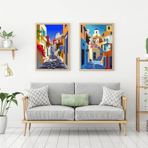 May include: Two framed illustrations of colorful buildings in a Greek village. The illustrations are set against a white wall with a couch and plants in the foreground.