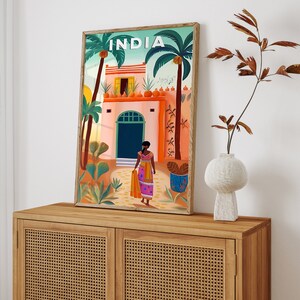 May include: A colorful illustration of a building in India with a woman in a pink and orange sari standing in front of it. The building has a green door and a red roof. The illustration is framed in a wooden frame.