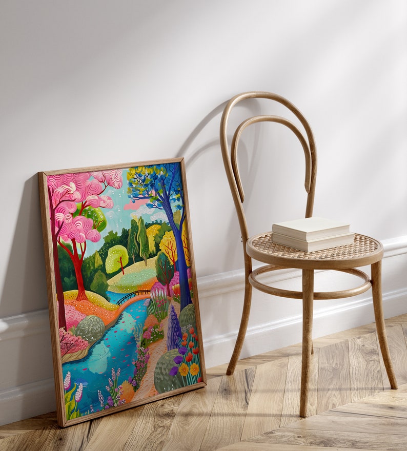 May include: A framed art print depicts a colorful landscape with a river, trees, and flowers. The artwork features vibrant hues of pink, blue, green, and yellow. A wooden chair with a woven seat and a stack of books sits nearby.