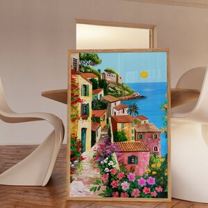 May include: A painting of a colorful Italian village with houses, flowers, and a view of the ocean. The sky is blue with a bright yellow sun. The painting is framed in wood.