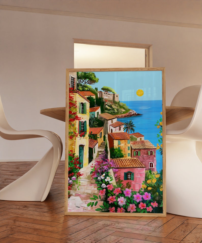 May include: A painting of a colorful Italian village with houses, flowers, and a view of the ocean. The sky is blue with a bright yellow sun. The painting is framed in wood.