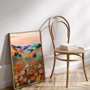 May include: A framed art print featuring a colorful landscape with rolling hills, trees, and a vibrant field of flowers in shades of pink, yellow, and orange. The artwork is displayed next to a wooden chair with a woven seat, with a stack of books on the seat.