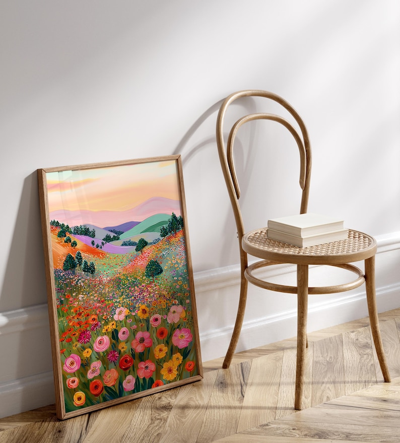 May include: A framed art print featuring a colorful landscape with rolling hills, trees, and a vibrant field of flowers in shades of pink, yellow, and orange. The artwork is displayed next to a wooden chair with a woven seat, with a stack of books on the seat.