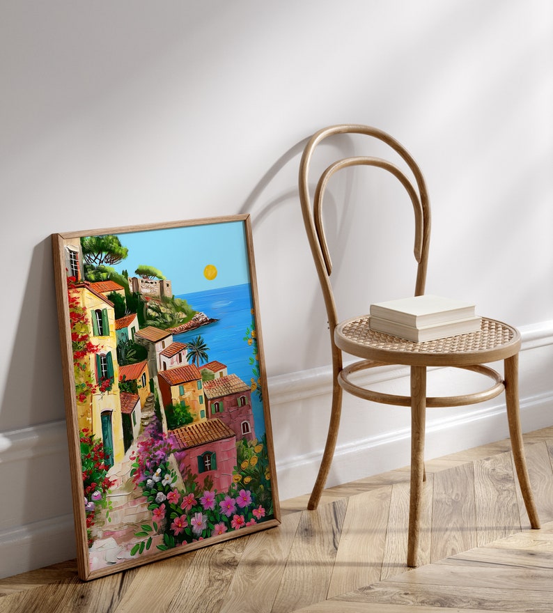 May include: A framed painting of a colorful Italian village with a blue ocean in the background. The painting is in a wooden frame and is leaning against a white wall.