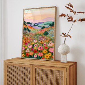 May include: A framed art print depicting a vibrant landscape with colorful wildflowers in the foreground. The artwork features rolling hills, trees, and a sunset sky with shades of pink, orange, and purple. The frame is a light brown color.