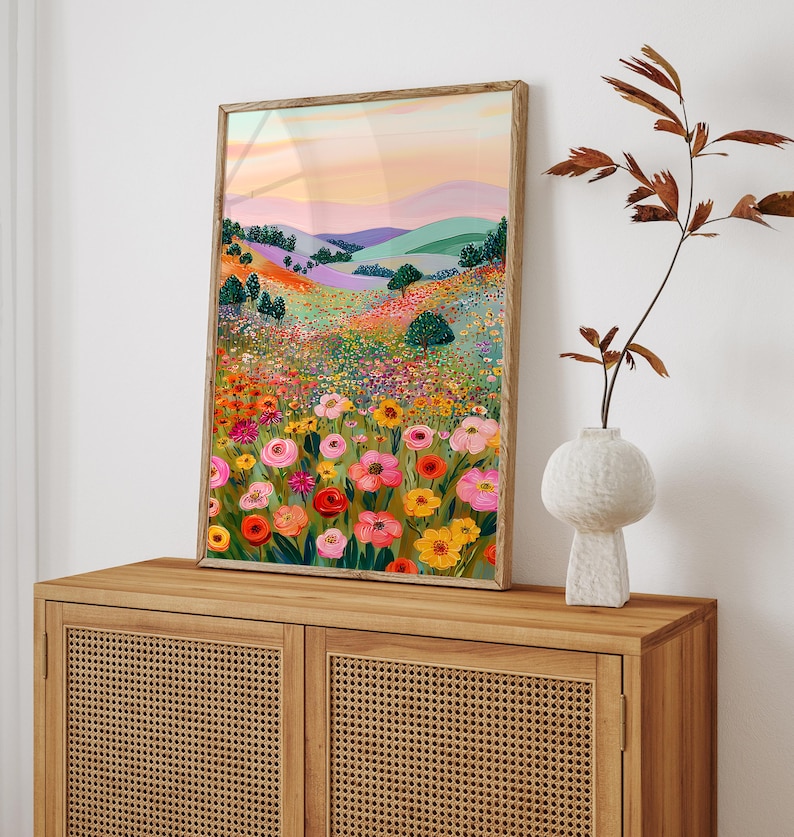 May include: A framed art print depicting a vibrant landscape with colorful wildflowers in the foreground. The artwork features rolling hills, trees, and a sunset sky with shades of pink, orange, and purple. The frame is a light brown color.