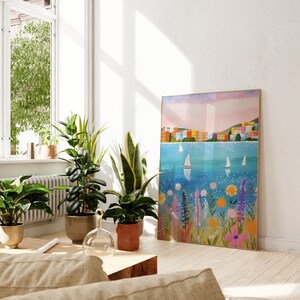 May include: A colorful painting of a seaside town with sailboats on a blue body of water. The town is made up of colorful buildings with white trim. The painting is framed in a light wood frame and is hanging on a white wall.