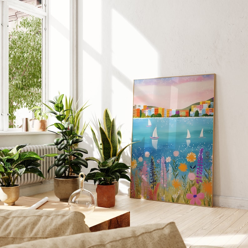 May include: A colorful painting of a seaside town with sailboats on a blue body of water. The town is made up of colorful buildings with white trim. The painting is framed in a light wood frame and is hanging on a white wall.