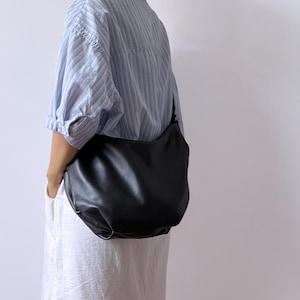 Può includere: Una borsa a tracolla in pelle nera dal design curvo. La borsa è indossata sulla spalla. La persona indossa una camicia a righe azzurre e un vestito bianco. La borsa ha un design semplice e minimalista.