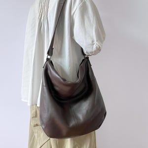 Könnte beinhalten: Eine dunkelbraune Leder-Umhängetasche mit langem Riemen. Die Tasche hat eine abgerundete Form und eine strukturierte Oberfläche. Der Riemen ist hellbraun. Die Tasche wird über der Schulter getragen.