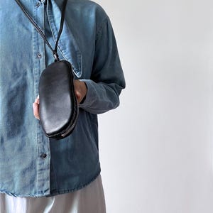 May include: A black leather crossbody bag with a rounded shape and a zipper closure. The bag is suspended from a thin black strap. The bag is held by a person wearing a denim shirt.