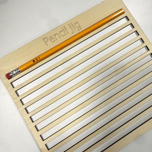 May include: A light-colored wooden pencil jig featuring a series of parallel horizontal cutouts and the text 'Pencil Jig' engraved on the top surface. A standard yellow HB pencil with a pink eraser is positioned horizontally across the top.