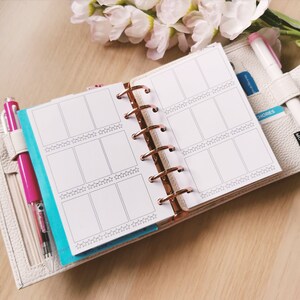 Puede incluir: Una agenda blanca con una encuadernación en espiral de color oro rosa. La agenda tiene una página con una cuadrícula de casillas, cada una con una fila de cinco estrellas en la parte inferior. La página está etiquetada como "CHORES".