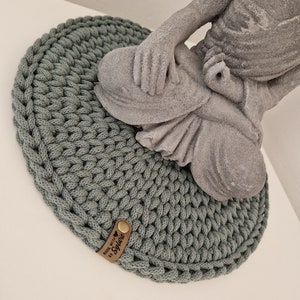 Könnte beinhalten: Ein runder, salbeigrüner Häkeluntersetzer mit einem braunen Lederanhänger, auf dem "Made with ♥ by Sophie" steht. Eine graue Beton-Buddha-Statue steht auf dem Untersetzer.