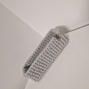 Könnte beinhalten: Ein hellgrauer, rechteckiger Häkelkorb mit gewebter Struktur. Ein kleines braunes Lederetikett mit einem Knopf ist an der Seite befestigt. Der Korb ist leer und steht auf einer weißen Oberfläche.