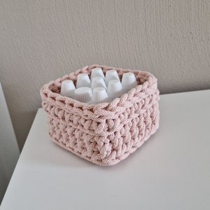 Könnte beinhalten: Ein rosa gehäkelter Korb gefüllt mit weißen Plastikflaschen. Der Korb ist aus dickem, strukturiertem Garn gefertigt und hat eine quadratische Form.