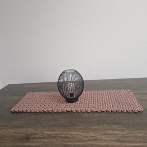 Könnte beinhalten: Eine schwarze Tischlampe aus Metall mit einem runden Käfigschirm steht auf einem rosa gehäkelten Tischläufer.