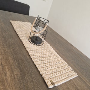 Könnte beinhalten: Ein beigefarbener gehäkelter Tischläufer mit einer schwarzen Metallkäfig-Leuchte darauf. Der Tischläufer liegt auf einem dunkelbraunen Holztisch.