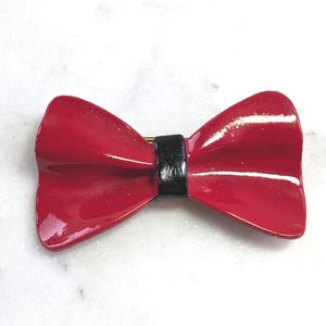 May include: A glossy red bow tie brooch with a black centre band. The brooch has a gold-coloured pin on the back. The bow tie is a classic accessory.
