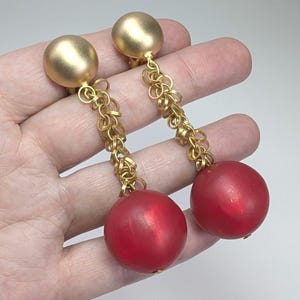 May include: A pair of gold-coloured clip-on earrings with a dangling design. Each earring features a gold sphere at the top, connected by a chain of small gold rings to a large, round, red bead. The earrings are shown against a light background.