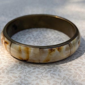May include: A circular bangle bracelet featuring a smooth, polished exterior composed of segmented, cream-coloured panels with natural brown mottled patterns. The interior of the bracelet is lined with a solid, dark metallic band that provides a contrast.