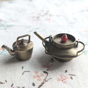 Puede incluir: Dos teteras de latón en miniatura, una con un pomo rojo en la tapa. Las teteras están sobre un fondo de tela blanca con un patrón floral.