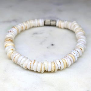 May include: A circular bracelet composed of numerous small, flat, disc-shaped white and cream-coloured shell beads. The beads are strung together in a continuous loop, featuring a single cylindrical silver-toned metal clasp connector at the top.