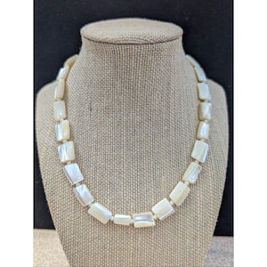 May include: A necklace made of rectangular, off-white mother-of-pearl beads. The beads have an iridescent sheen and are strung together to form a continuous loop. The necklace is displayed on a mannequin with a neutral tan background. The beads are rectangular.