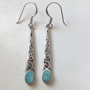 May include: A pair of silver dangle earrings with a turquoise stone set in each. The earrings have a delicate, filigree design.