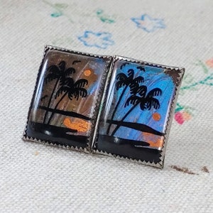 May include: A pair of silver cufflinks featuring a blue and brown butterfly wing design. The wings are set in a silver frame and depict a silhouette of palm trees and a sunset.