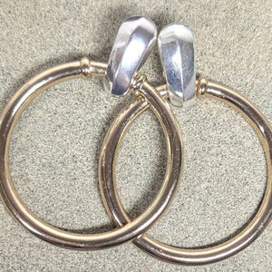 May include: A pair of gold-colored hoop earrings with silver-colored, geometric-shaped clasps. The earrings are circular and have a smooth, polished finish. The clasps are rectangular with a faceted design.