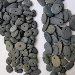May include: A collection of gray, drilled beach stones. The stones vary in size and shape, with a hole drilled through each one. The stones are a mix of light and dark gray, and are arranged in two piles against a white background. Ideal for jewelry making.