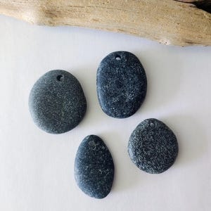May include: Four dark gray, smooth river stones with drilled holes for jewelry making or crafts. The stones vary in size and shape, with a natural, textured appearance. A piece of driftwood rests in the background.