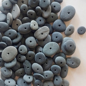 May include: A large pile of gray and blue smooth river rocks with holes drilled through the center. The rocks are various sizes and shapes, and they are perfect for crafting.